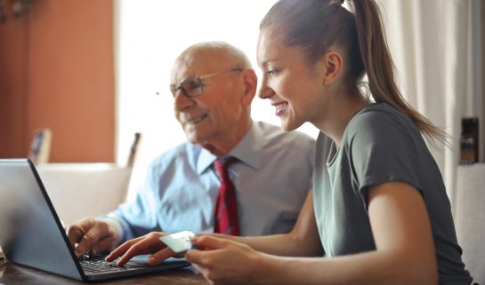 A young woman holding a credit card and an elderly man sit in front of a laptop