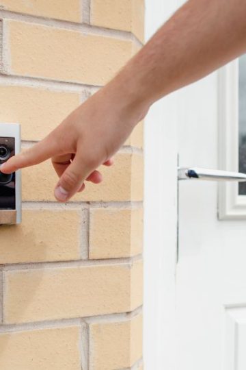 An image of a hand pressing a smart doorbell that is mounted on a brick wall next to a white door.